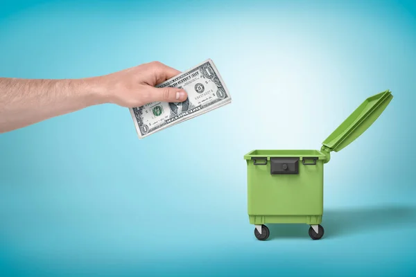 Male hand throwing bundle of money dollars into green trash bin on blue background