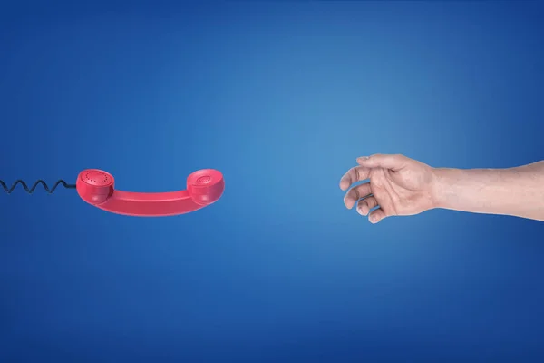 Un receptor de teléfono retro rojo cuelga frente a una mano masculina vacía sobre un fondo azul . —  Fotos de Stock