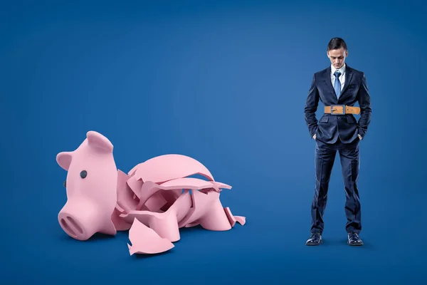 Zakenman, het dragen van strakke riem en gebroken piggy bank op blauwe achtergrond — Stockfoto