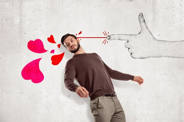 Young handsome man in casual clothes standing in half-turn near wall and leaning back as if from hand-drawn finger gun shooting pink hearts at him.