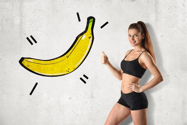 Young fit woman in black gym crop top and shorts standing in half-turn doing thumbs up pointing at drawing of big banana on wall.