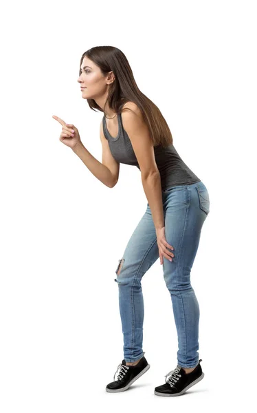 Seitenansicht einer jungen schönen Frau in grauem Top und blauer Jeans, die sich nach vorne beugt und auf etwas zeigt, mit Zeigefinger isoliert auf weißem Hintergrund. — Stockfoto