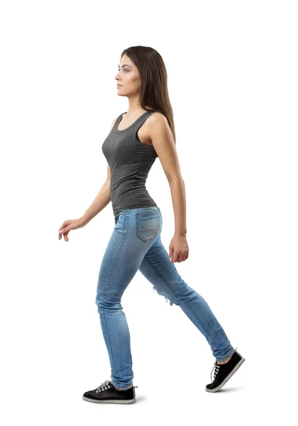 İleri beyaz arka plan üzerinde yürüyen genç kadın gri kolsuz üst ve fındık uzun siyah saçlı, mavi kot pantolon yan görünüm. — Stok fotoğraf