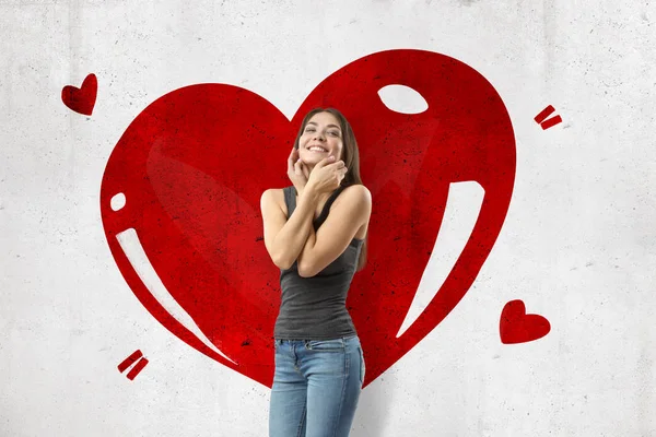 Jeune fille brune portant un jean décontracté et un t-shirt visage souriant et touchant avec un grand cœur rouge dessiné sur fond de mur blanc — Photo