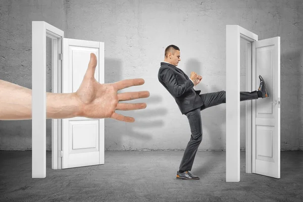 Businessman kicking a door and big hand appearing out of an open door on grey wall background