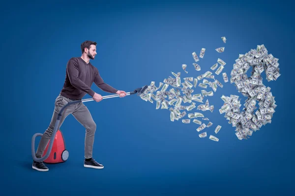 Zijaanzicht van de mens in casual kleding hoovering dollar en grote dollarteken met stofzuiger. — Stockfoto