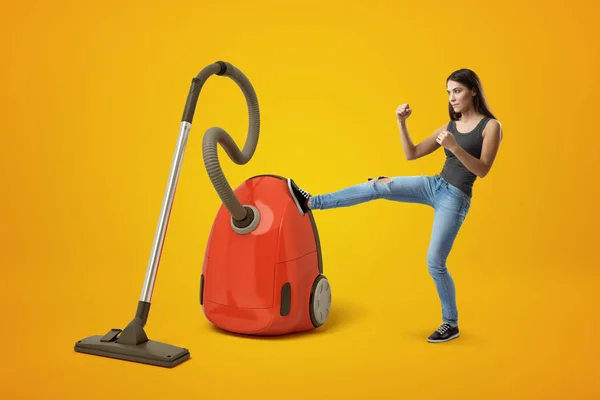 Mujer joven en pantalones vaqueros grises y azules, puños apretados y levantados, pateando gran aspiradora roja con pierna derecha sobre fondo amarillo . — Foto de Stock