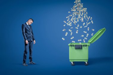 Üzgün işadamı yağmur içine düşen dolar ile açık dumspter yanında durduğunu aşağı bakıyor.