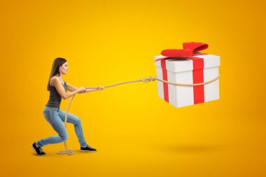 Side view of young woman standing with bent knees and pulling big gift box in air which she has lassoed, on yellow background. clipart