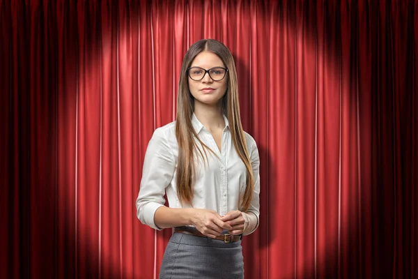 Young attractive woman in white office shirt, grey skirt and glasses, standing in spotlight against red stage curtain.