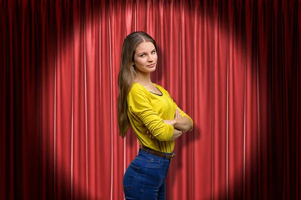 Side close-up of young woman in yellow jumper and blue jeans lit up by limelight standing with arms folded and looking at camera against red curtain.