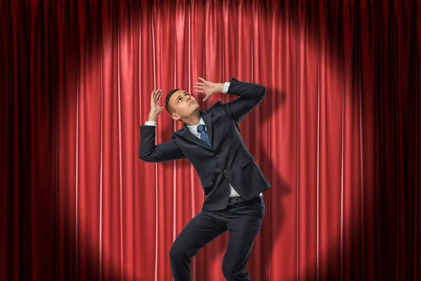 Businessman standing in spotlight near red stage curtain, leaning aside, looking up and protecting head with hands from something above and out of view.