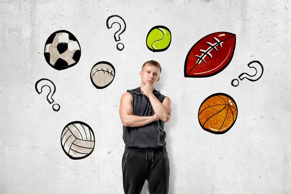 Vista frontal del joven con capucha sin mangas de pie con la mano frotando la barbilla en la pared con dibujos de diferentes pelotas deportivas y signos de interrogación entre ellos . — Foto de Stock