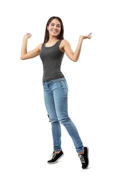 Young smiling woman in sleeveless gray top and blue jeans standing on tiptoes with arms bent and raised to sides isolated on white background. — Stock Photo, Image
