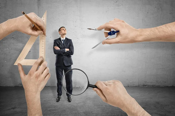 Businessman, arms folded, standing and looking up, and four big hands around measuring him with magnifying glass, drawing compass and triangle. — Stock Photo, Image