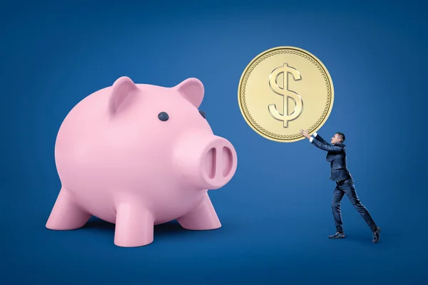 Tiny businessman holding big golden coin and big pink piggy bank on blue background — Stockfoto