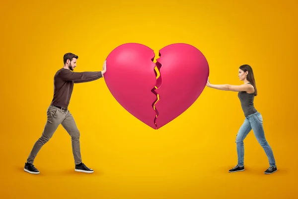 Young man and woman wearing casual clothes holding pink broken heart together on yellow background — Φωτογραφία Αρχείου