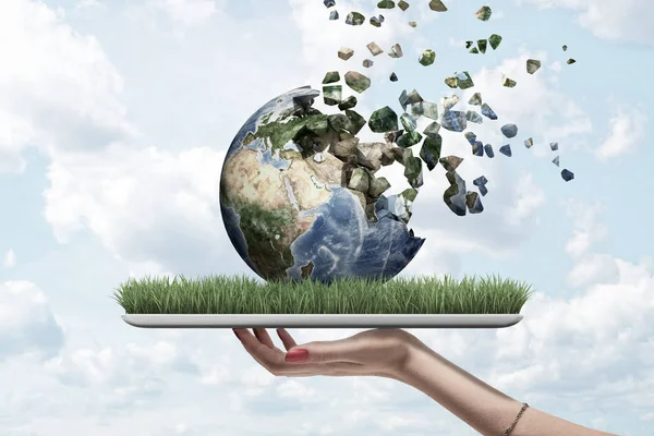 Side view of womans hand holding tablet with green grass on screen and Earth globe on top which is dissolving into pieces that float away into sky.