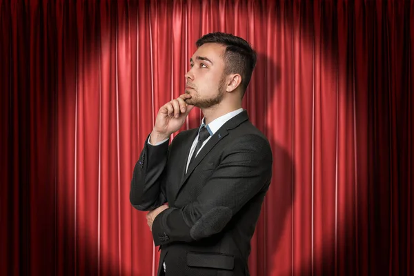 Nahaufnahme eines jungen Geschäftsmannes, der in halber Drehung mit der Hand am Kinn vor dem roten Bühnenvorhang steht und wegschaut. — Stockfoto