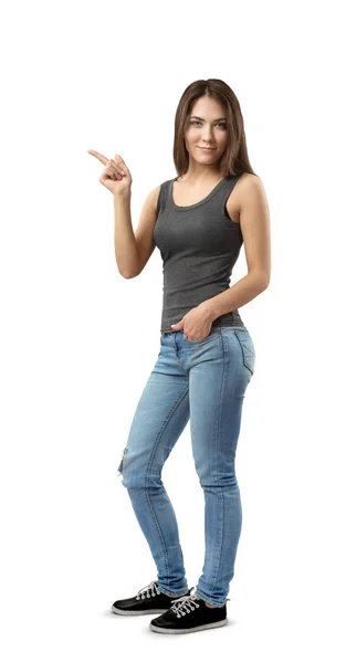 Junge Frau in ärmellosem Oberteil und Jeans, die in halber Drehung steht, mit der linken Hand in der Tasche und der rechten Hand, die seitlich isoliert auf weißem Hintergrund zeigt. — Stockfoto