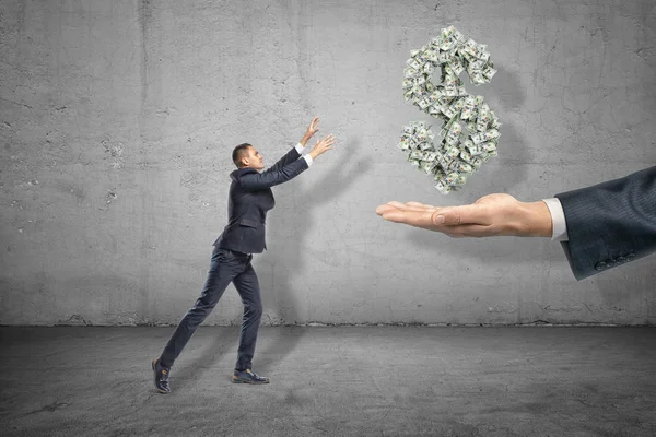 Вид сбоку миниатюрного бизнесмена, держащегося за руки, чтобы схватить символ доллара, образованный из многих банкнот, подвешенных в воздухе над большой рукой . — стоковое фото