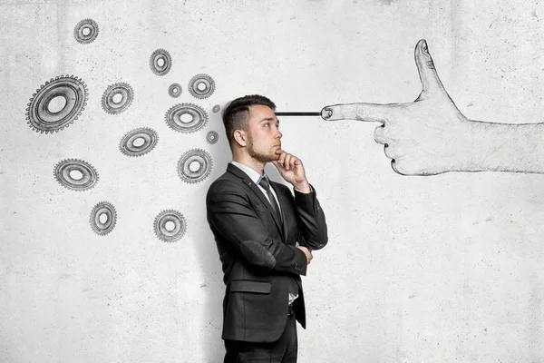 Thinking young businessman with a hand shooting gesture and gear wheels drawn on white wall background