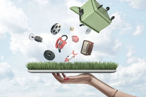 Side view of womans hand holding tablet with green grass on screen and green dumpster upside down in air out of which misc objects are falling.