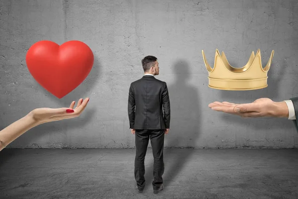 Young businessman standing between two big hands holding red heart and golden crown on grey wall background. background — 스톡 사진