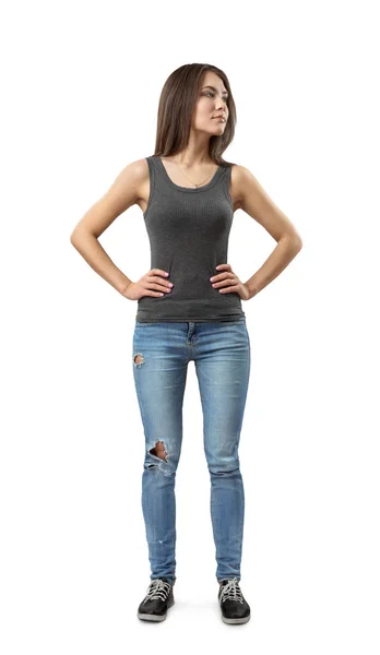 Front bild av ung attraktiv kvinna i grå ärmlös topp och blå jeans stående och ser bort med händerna på höfter isolerade på vit bakgrund. — Stockfoto