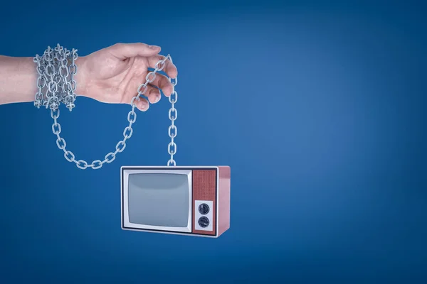 Close-up of mans hand with metal chain around wrist holding retro TV set hanging on the other end of chain.