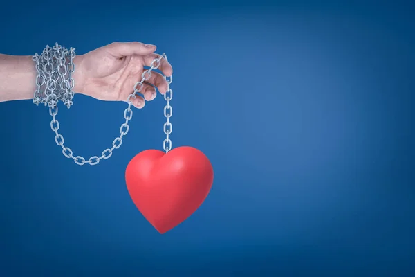 Close-up de mão de homem com corrente de metal em torno do pulso segurando coração valentine vermelho pendurado na outra extremidade da cadeia . — Fotografia de Stock