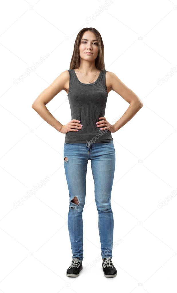 Front view of young attractive woman in gray sleeveless top and blue jeans standing with arms on waist isolated on white background.