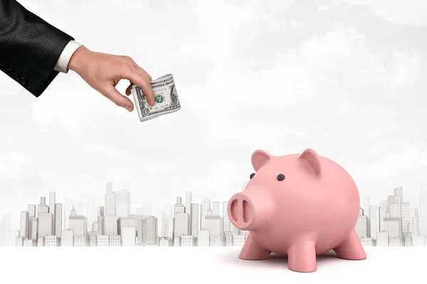 Mannelijke hand houden geld dollars met roze Piggy Bank op witte stad wolkenkrabbers achtergrond — Stockfoto