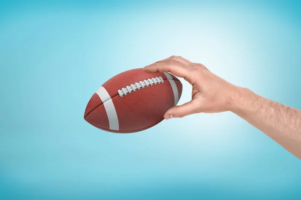 Mans mano sosteniendo la pelota para el fútbol americano sobre fondo degradado azul claro . — Foto de Stock