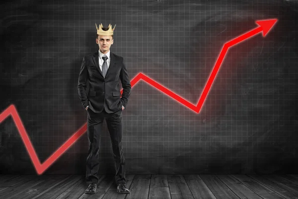 Full length front view of businessman wearing crown, standing hands in pockets, with red graph arrow going up behind him at black graph-ruled wall.