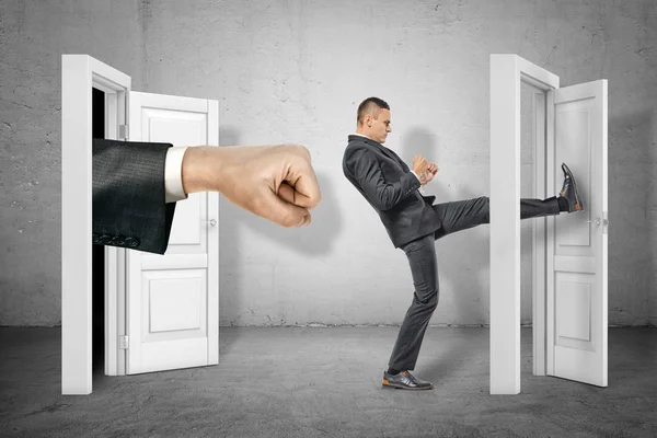 Businessman kicking door and big stretched fist appearing out of open door on grey wall background — Stock Photo, Image