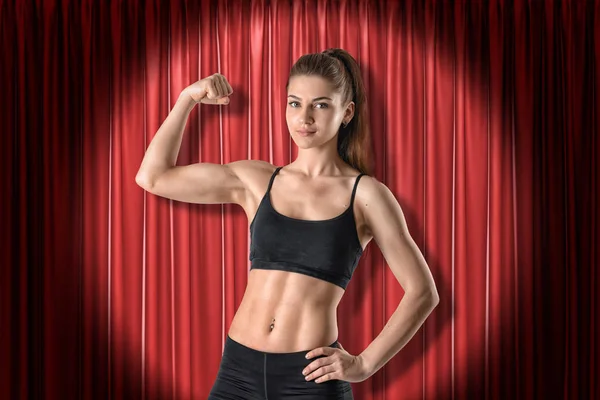 Fiatal sportos lány fekete sport ruházat mutatja bicepsz piros színpad függöny háttér — Stock Fotó