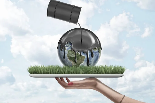 Female hand holding earth globe covered with black thick liquid from black oil barrel on green grass model on blue sky background