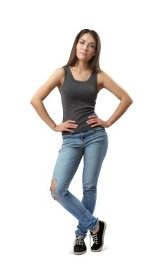 Young woman in gray sleeveless top and blue jeans standing with hands on hips and one leg in front of the other isolated on white background.