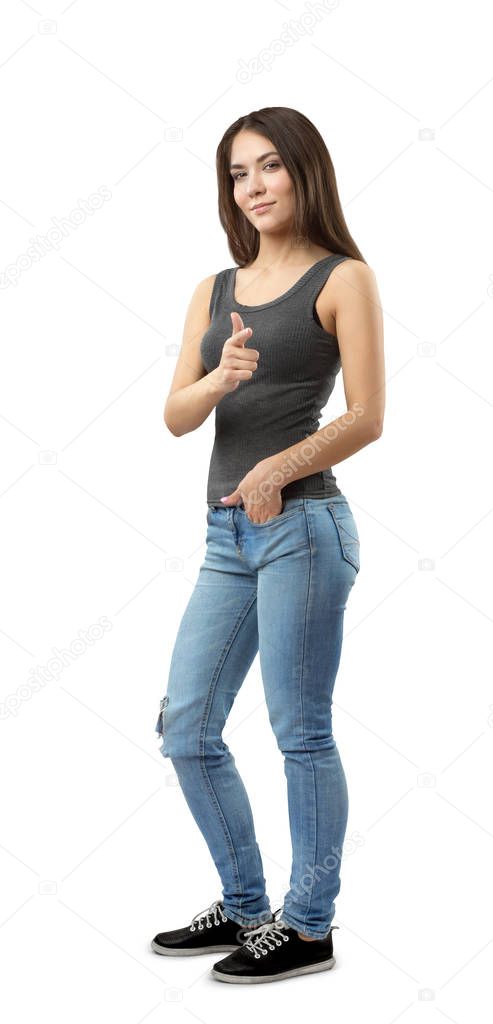 Young woman in sleeveless top and jeans standing in half-turn with left hand in pocket and right hand pointing at viewer isolated on white background.