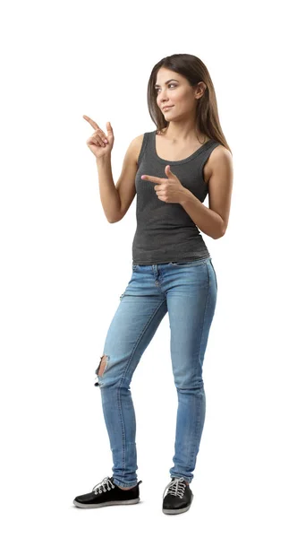 Young woman in sleeveless top and jeans standing in half-turn with both hands bent and index fingers pointing sideward isolated on white background. — 스톡 사진