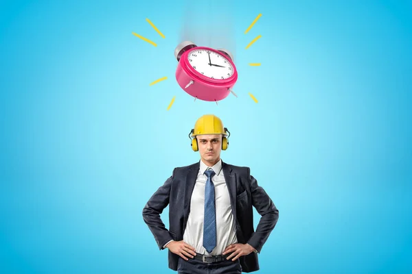 Front waist-deep image of businessman standing, hands on hips, wearing yellow hard hat with big pink alarm clock ringing in air above.