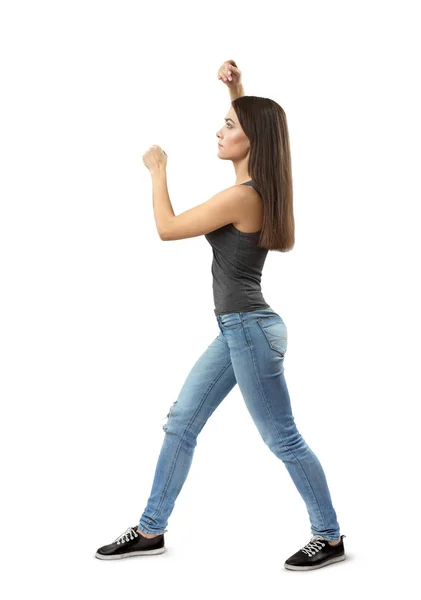 Side view of young woman in gray top and blue jeans standing, fist before face. other arm above head, right foot forward isolated on white background.