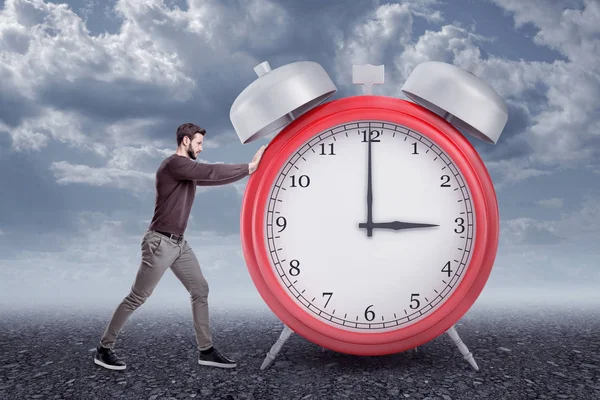 Vista lateral del joven barbudo con ropa casual empujando un gran despertador rojo, parado afuera bajo un cielo gris nublado . —  Fotos de Stock