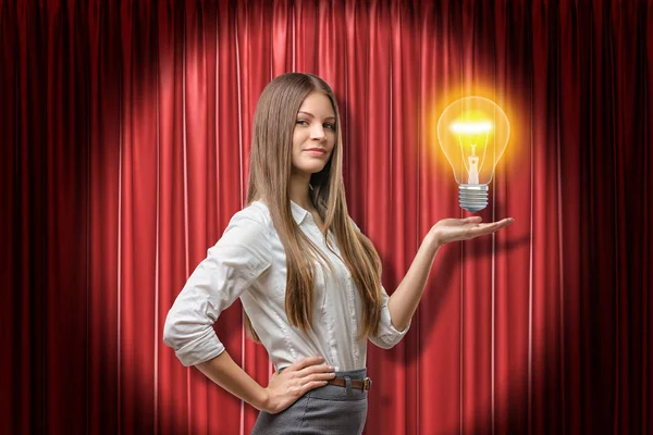 Jong brunette zakenvrouw holding neon geel gloeilamp op rood podium gordijnen achtergrond — Stockfoto