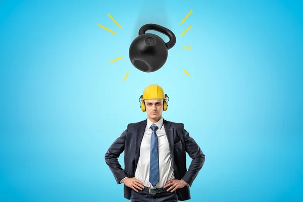 Crop kép üzletember sárga kemény kalap fülvédők, állva kezét a csípő, és a nagy nehéz kettlebell zuhan rá a fejét. — Stock Fotó