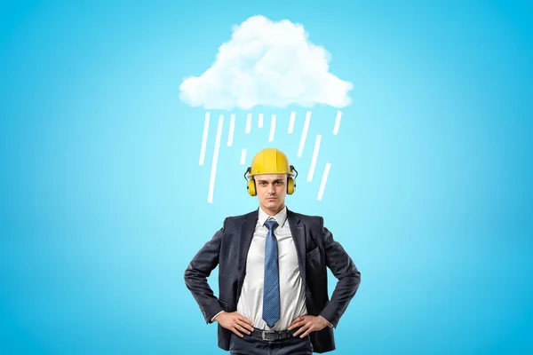 Front crop image of businessman in yellow hard hat with ear defenders, standing with hands on hips under white raining cloud.