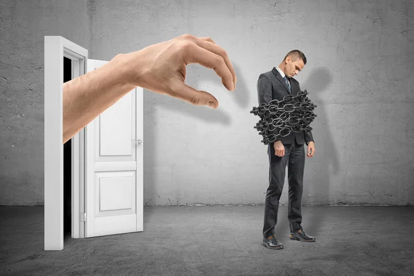 Volledige lengte zijaanzicht van droevige kleine zakenman die zich met kettingen rond zijn lichaam en reusachtige hand bevindt die van deur achter hem voortkomen. — Stockfoto