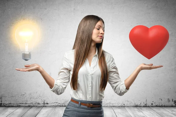 Jeune femme d'affaires tenant coeur rouge et ampoule jaune dans ses mains sur fond de mur gris — Photo