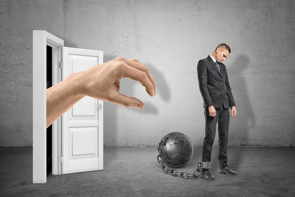 Big hand reaching through white doorway to catch young sad businessman who is chained to metal ball on grey wall background